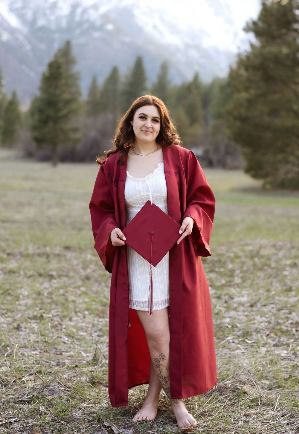Standing barefoot in red graduation gown holding decorated cap in mountain setting.
