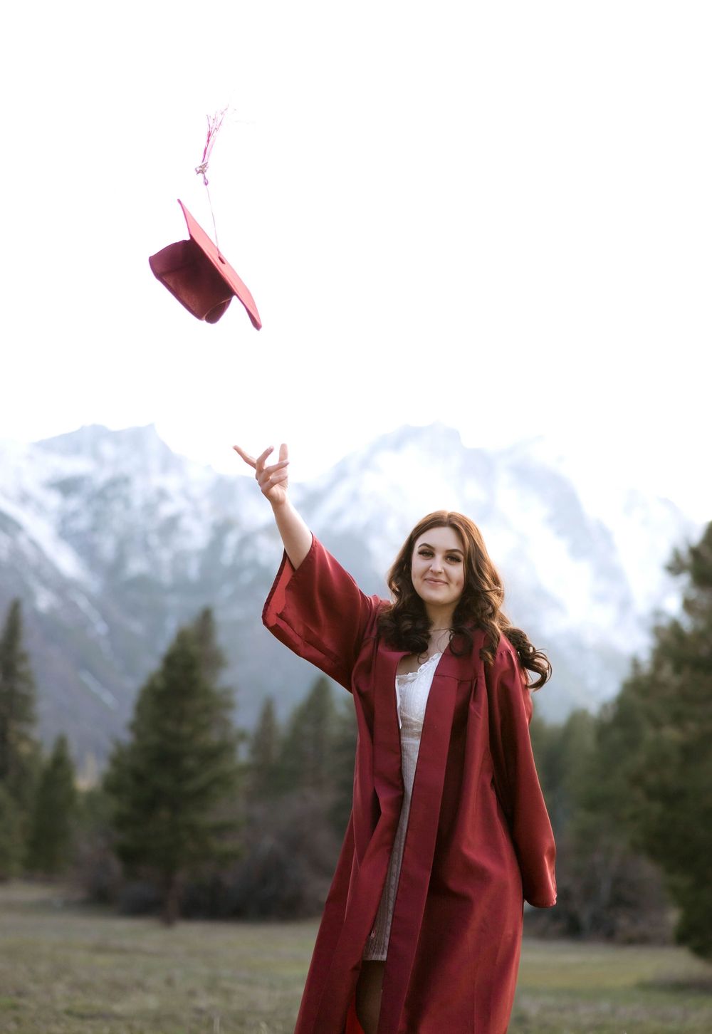 Graduation photo throwing cap in air wearing red gown with mountain backdrop.