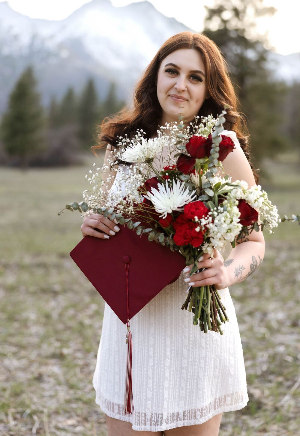 Holding graduation cap and floral bouquet in white dress against mountain landscape.
