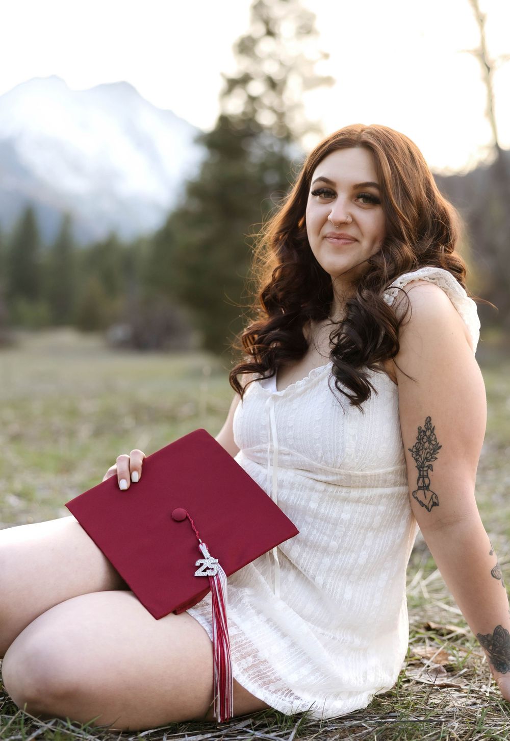 Sitting with graduation cap in white dress showing tattoo with mountains in background.