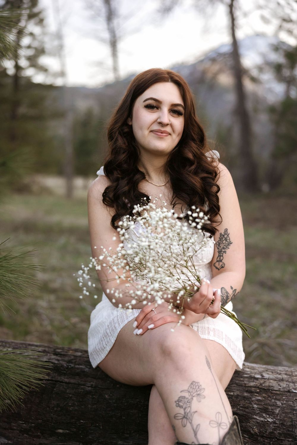 Sitting on log holding baby's breath flowers in white dress with mountain scenery.