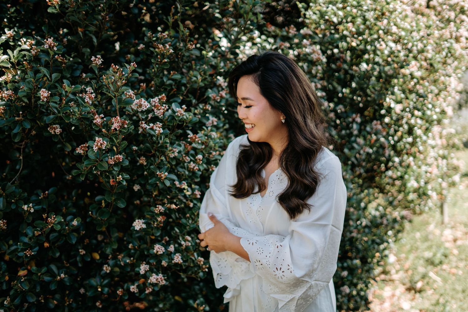 A person in a white lace dress poses outdoors against a lush green foliage background in a romantic photoshoot setting.