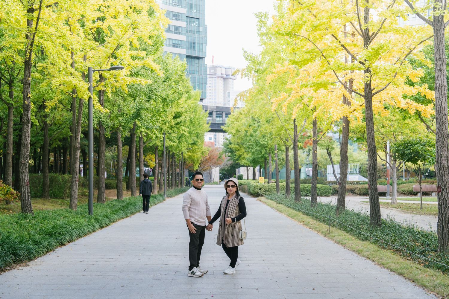 Two people walking on a tree-lined path with yellow autumn foliage on a sunny day.