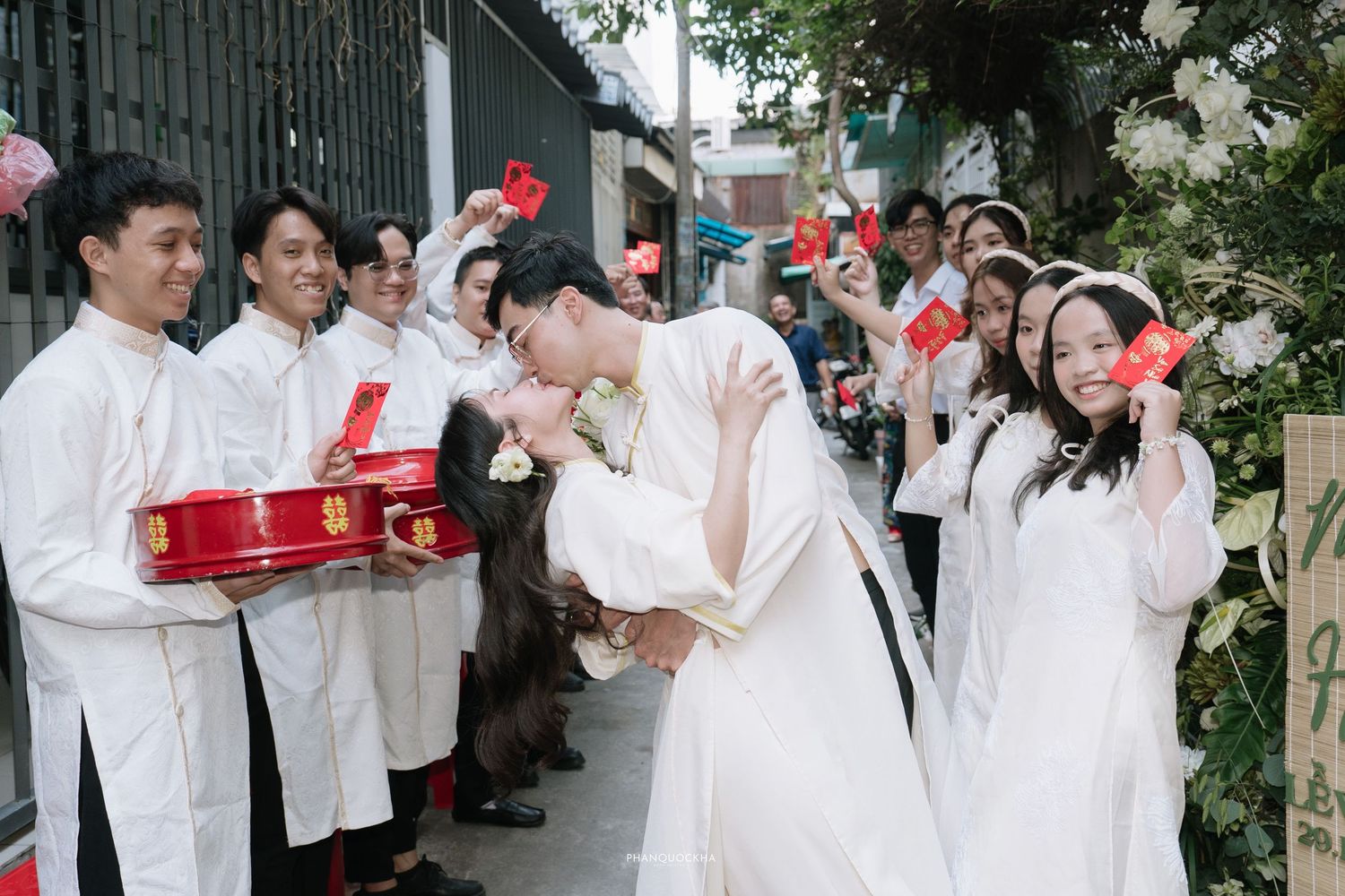 Vietnamese bride and groom kiss during traditional wedding procession with guests in white ao dai nearby.