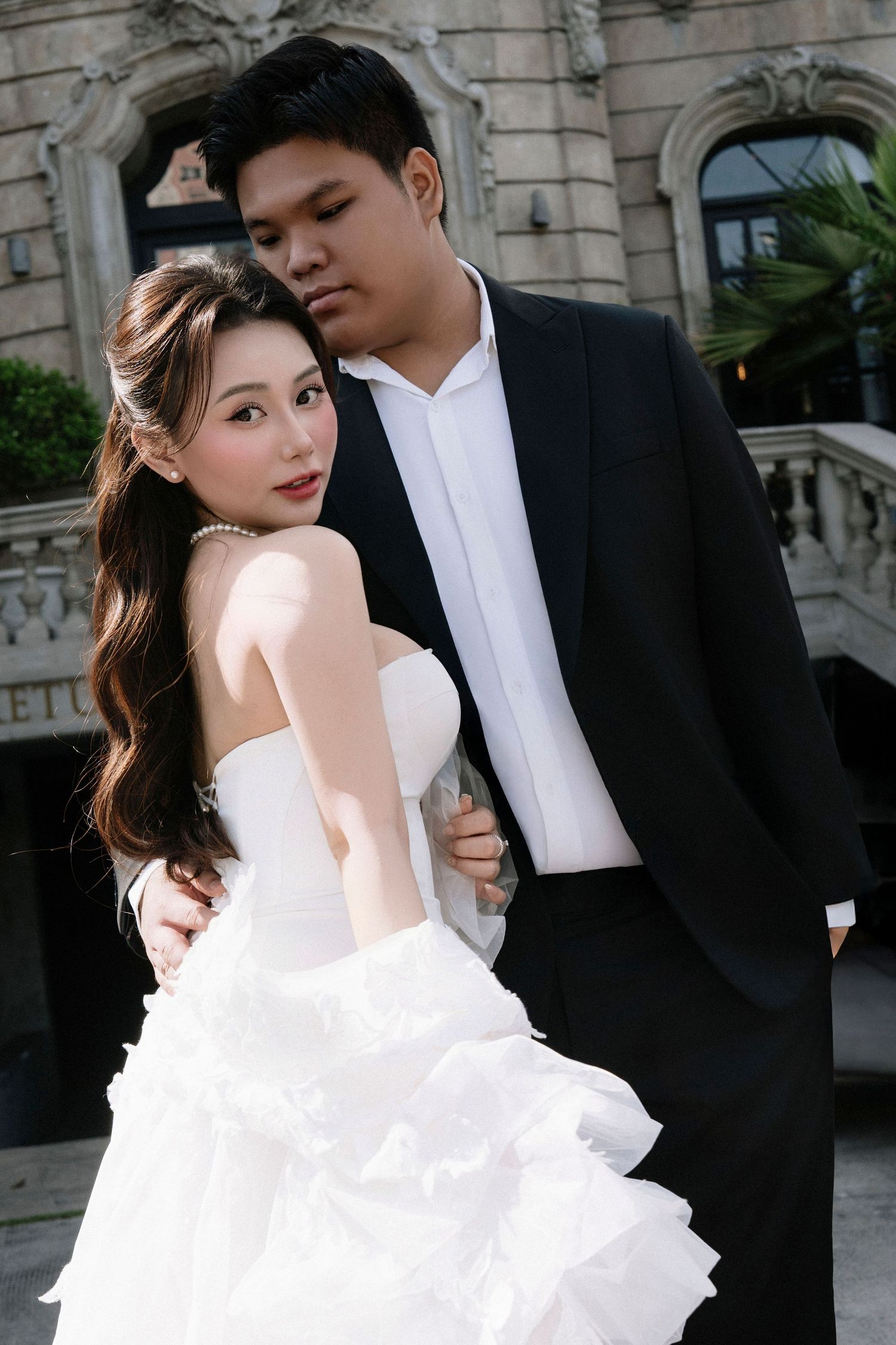 Asian couple in wedding attire embrace romantically, bride in white gown and groom in black tuxedo.