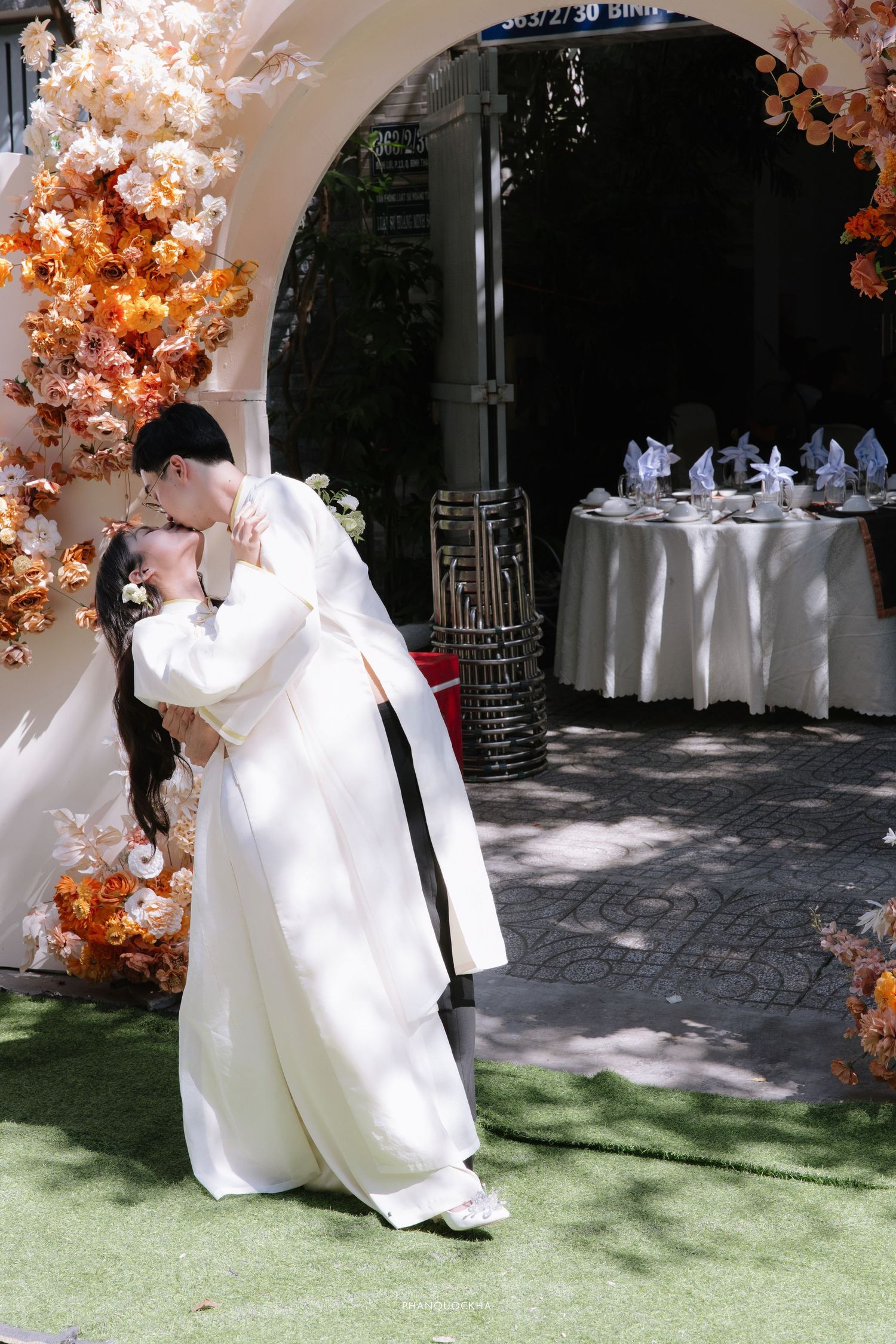 Vietnamese couple sharing a romantic kiss under floral arch at outdoor wedding ceremony in traditional ao dai attire.