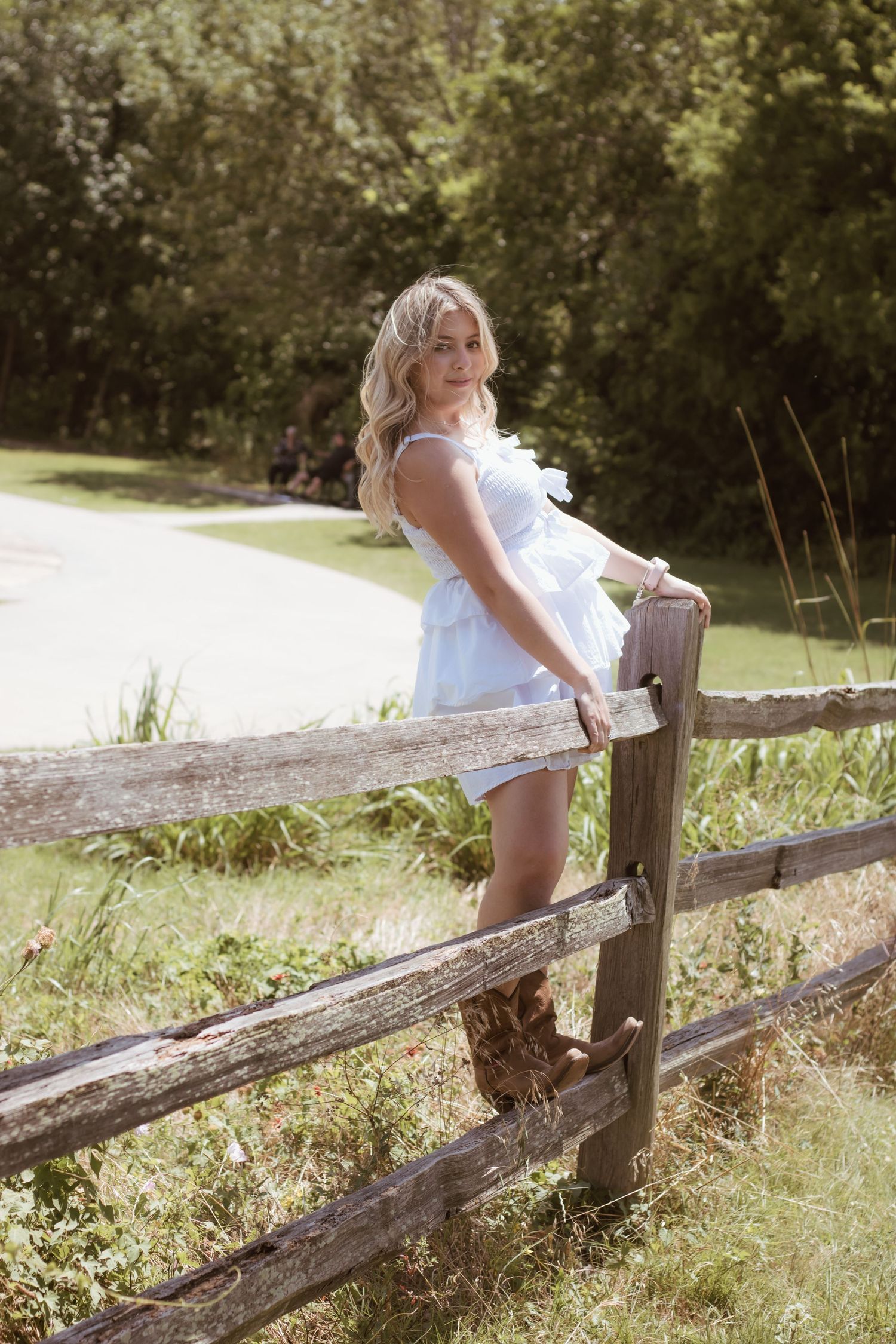 Woman in white dress poses casually on rustic wooden fence in summer setting.