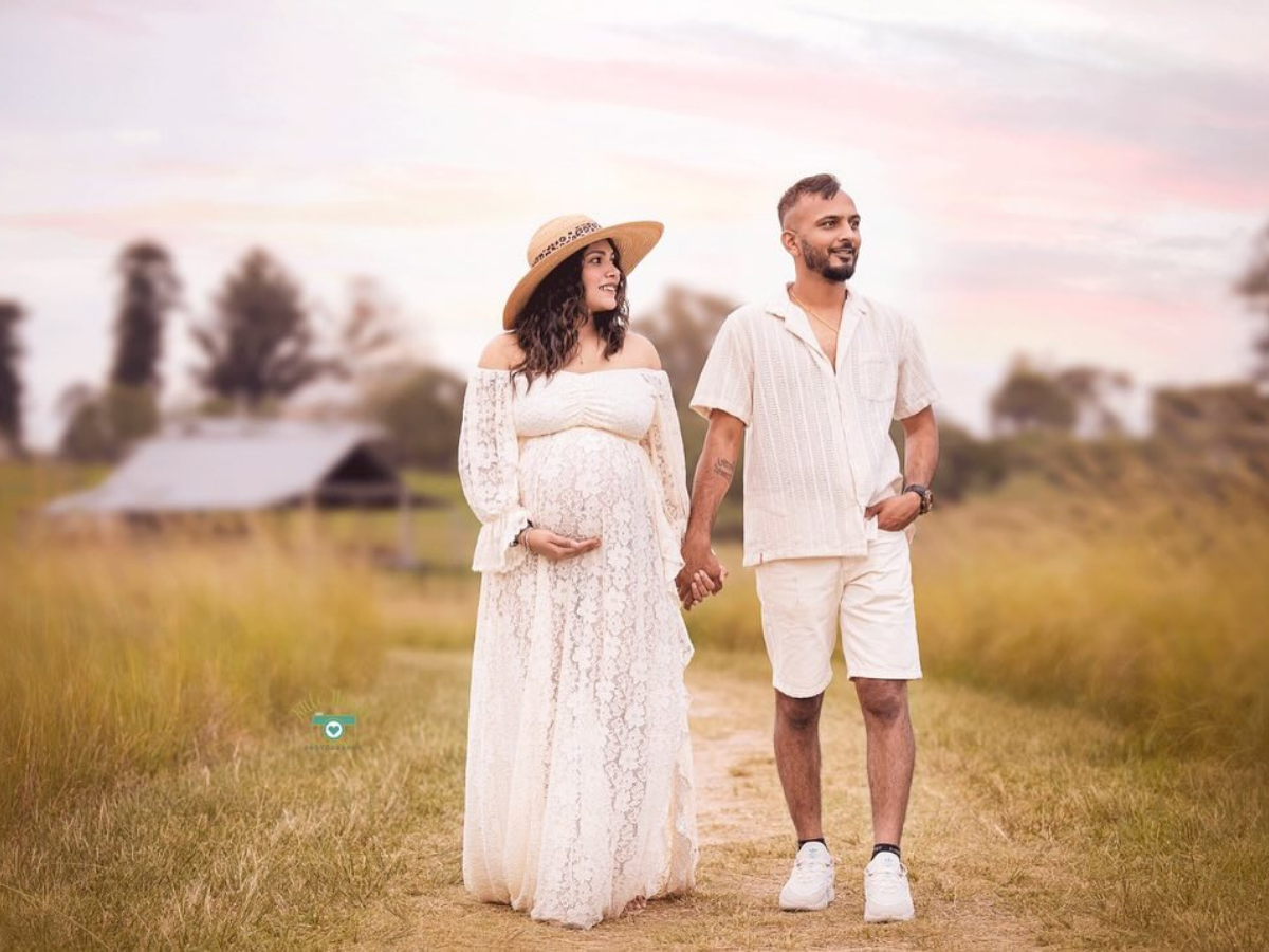 A romantic couple in white outfits walks together through a sun-lit grassy field at sunset.