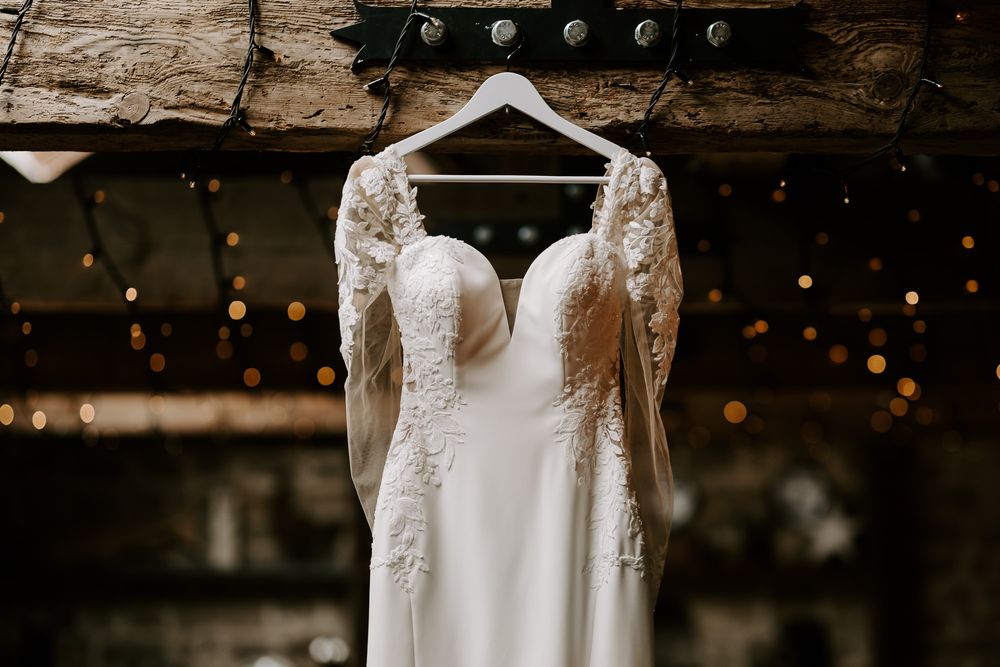 A wedding dress hangs elegantly near a window, showcasing delicate lace details and beading in natural light.