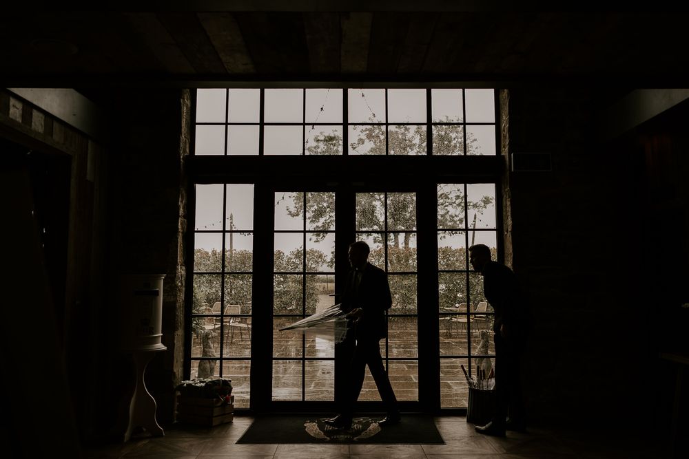 Silhouettes of a wedding party stand in front of large industrial windows in a moody, dark venue.