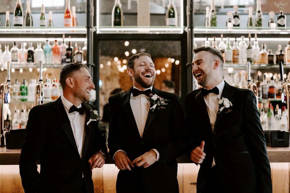 Group of groomsmen in formal attire laughing and socializing at an indoor reception venue.