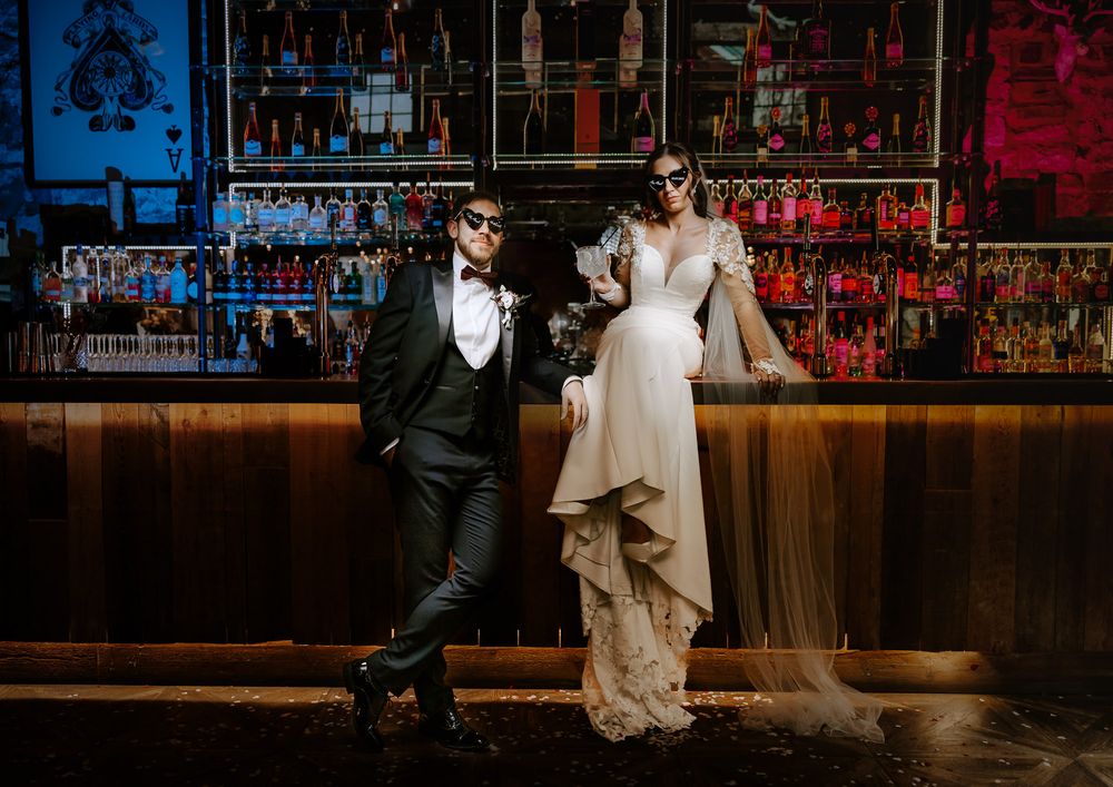 A couple poses at an elegant bar with dramatic red and blue lighting while dressed in formal wedding attire.