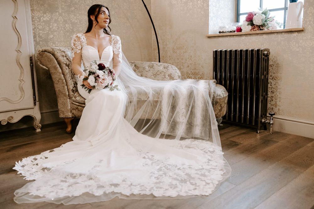 Elegant bride in long-sleeved lace wedding gown with flowing veil sits on vintage settee holding bouquet in classic interior.