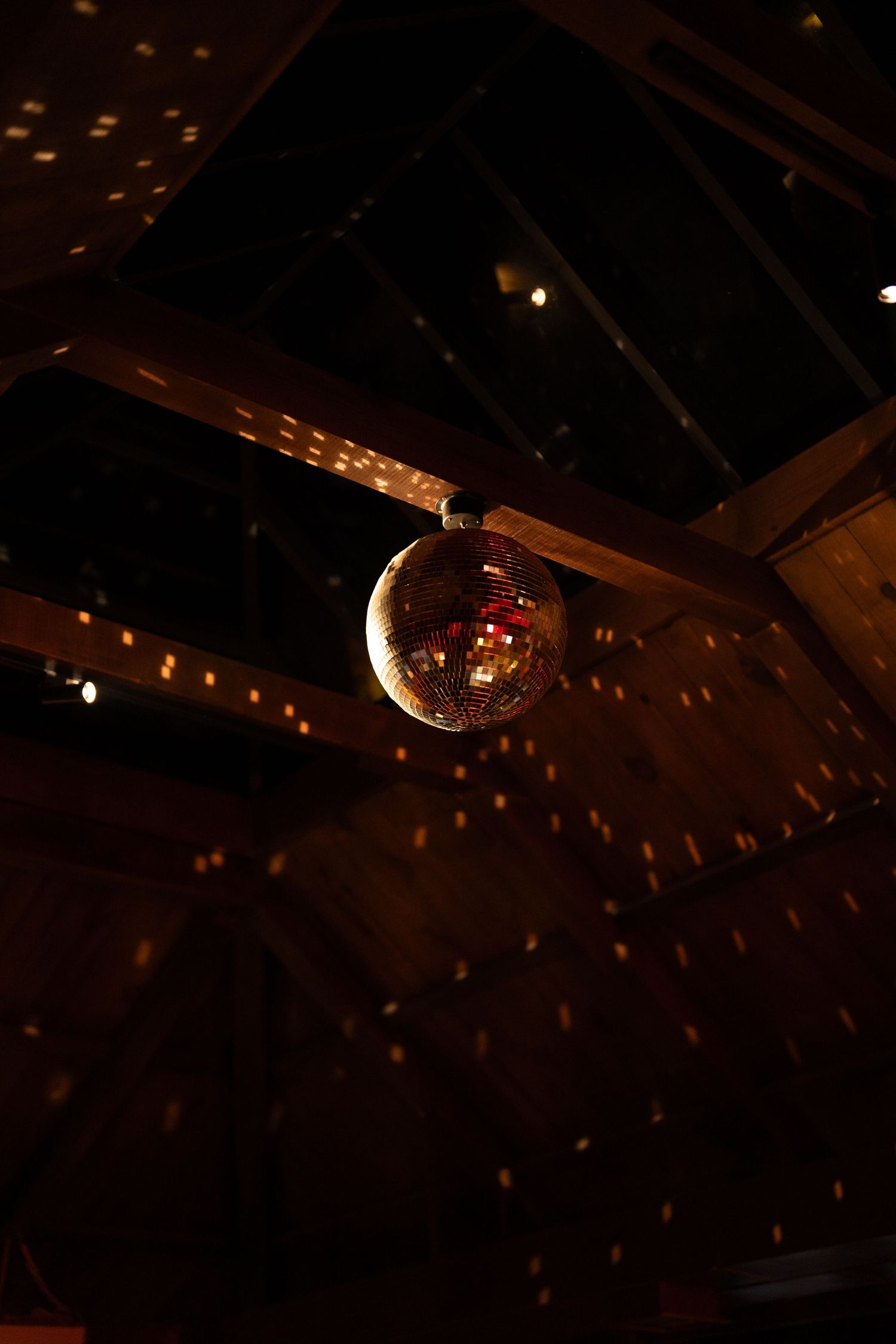 Glowing disco mirror ball hanging from dark wooden ceiling beams with warm scattered light reflections.