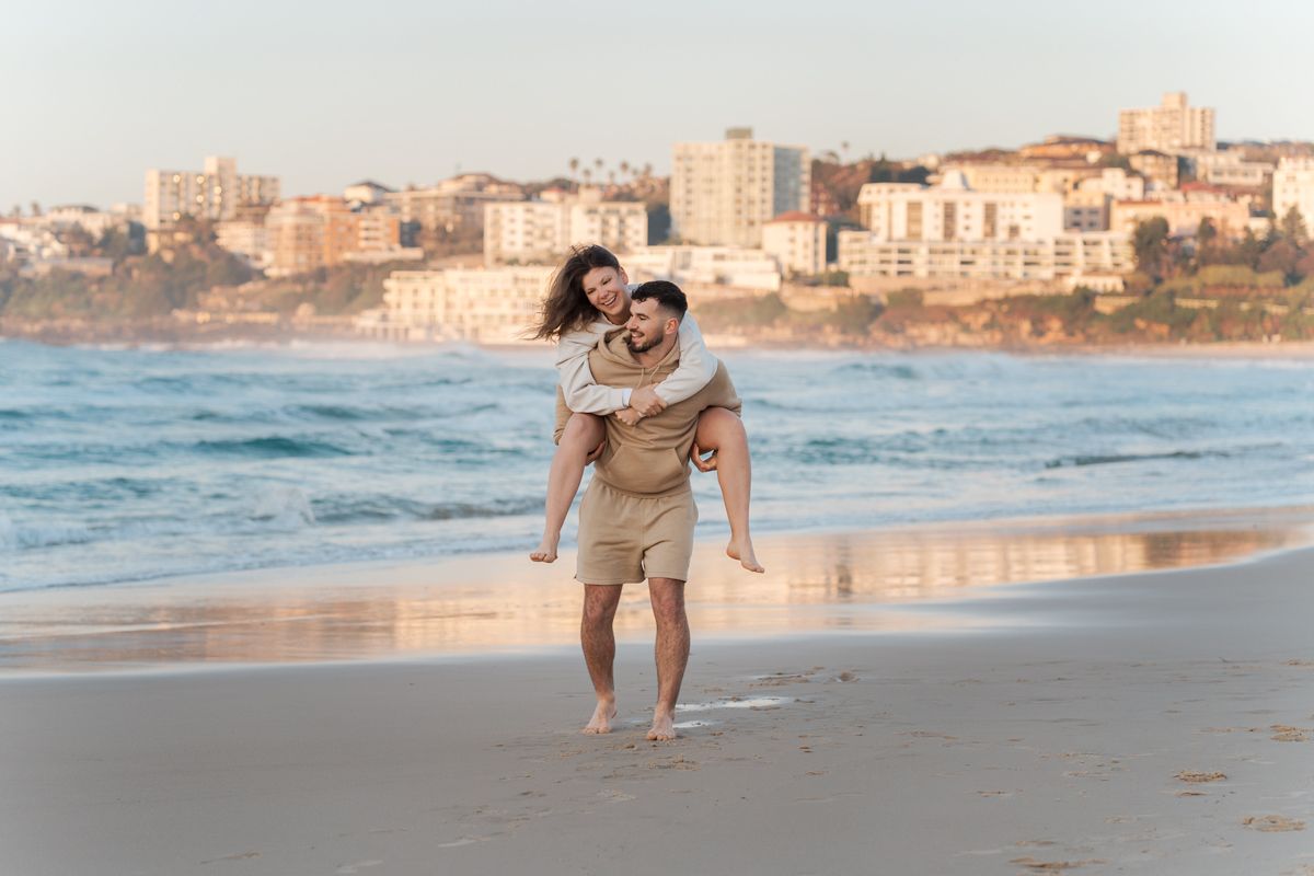 A couple enjoys a playful piggyback ride along Bondi beach at sunrise with coastal buildings in the background during a Sydney Photoshoot