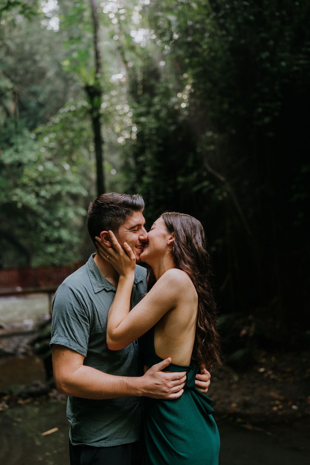 A romantic couple in matching green outfits share intimate moments by a waterfall in a lush forest setting.