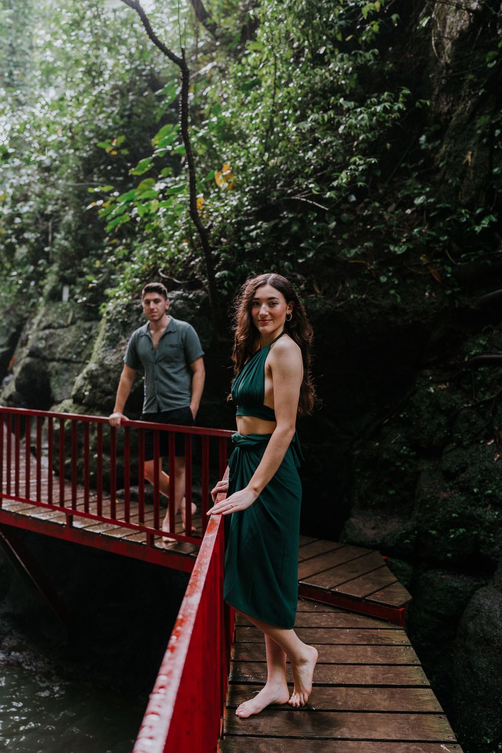 Couple in green attire exploring a red-railed wooden walkway in a forest setting.