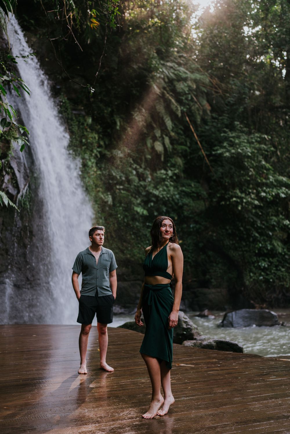 Couple in casual attire standing on a wooden deck with a waterfall view.