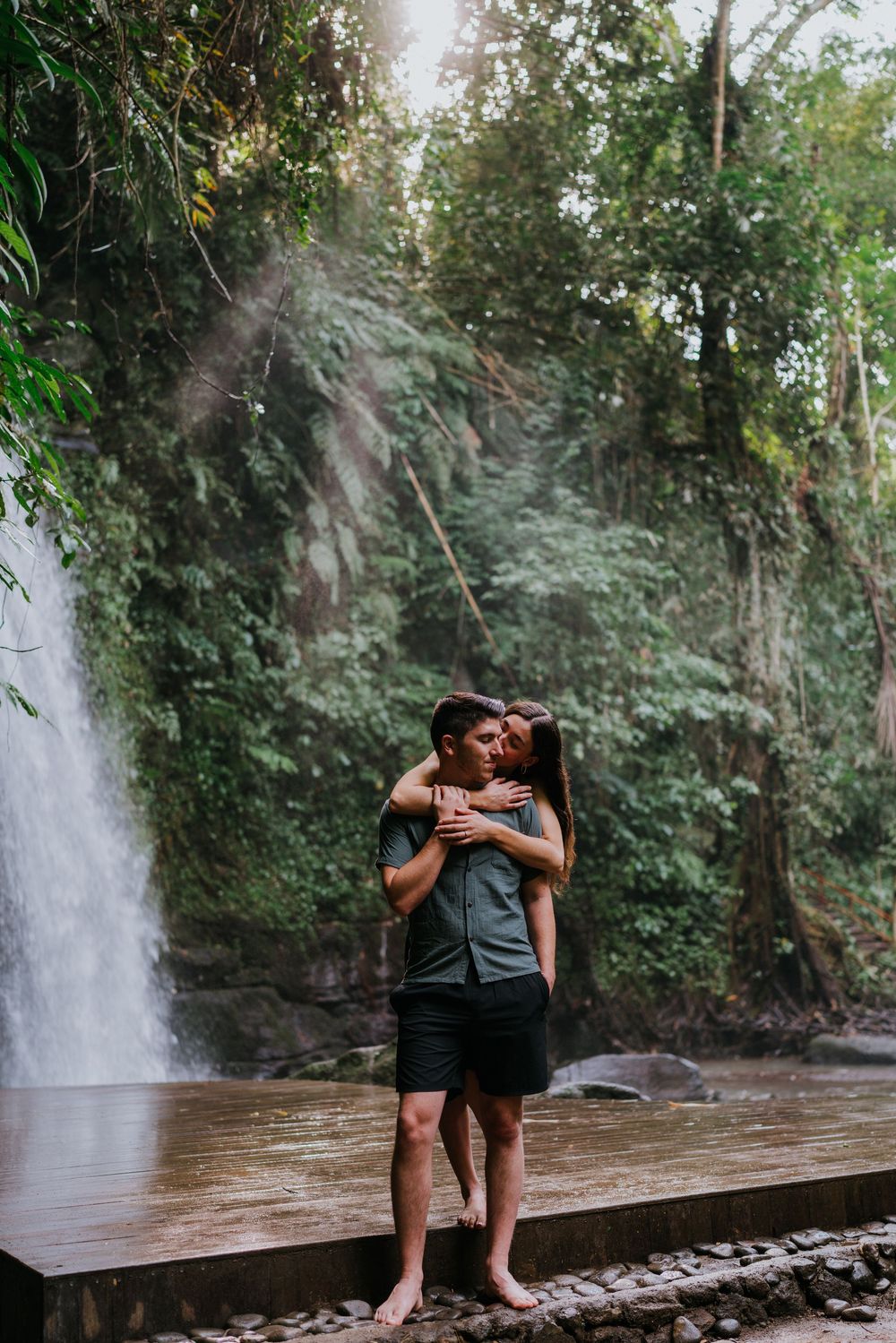 Two people embrace on a wooden boardwalk with a misty waterfall creating a dreamy atmosphere.