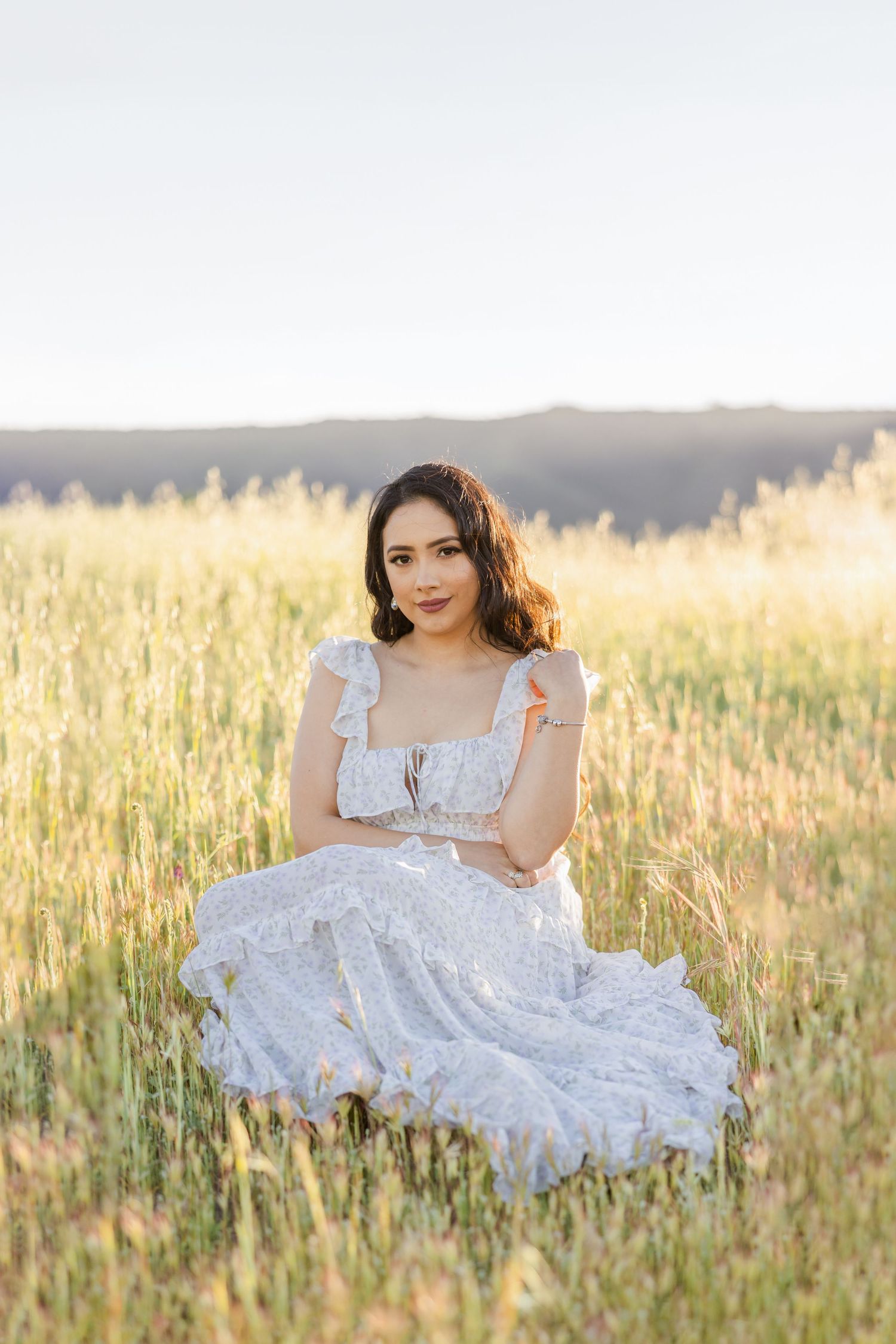 Someone in a white lace dress sits gracefully in a sunlit field of golden wheat at sunset.