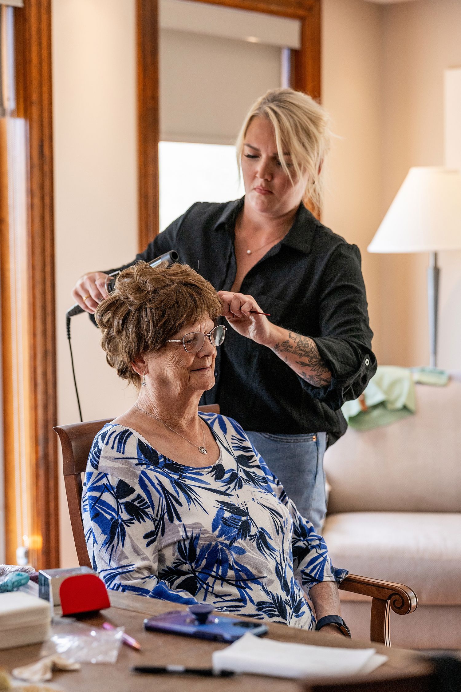 A hairstylist styles a client's hair in a bright salon setting with warm wood trim and natural lighting.
