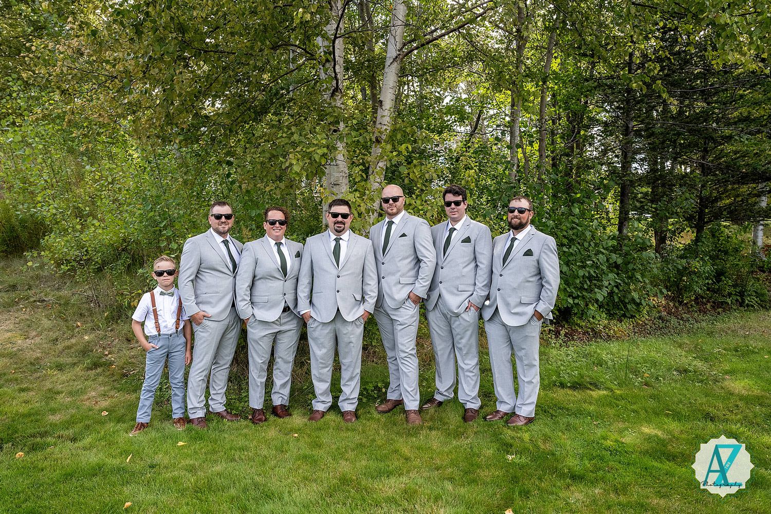 A wedding party in matching gray suits and suspenders poses together in a lush green outdoor garden setting.