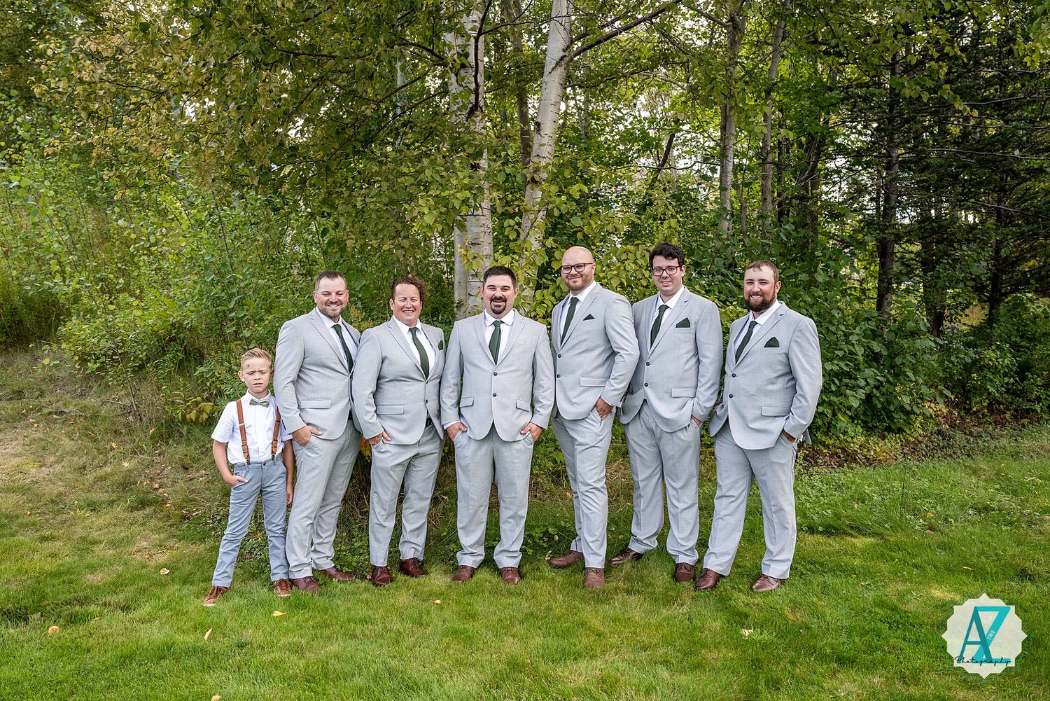 Wedding party wearing light gray suits stands in line against vibrant green foliage backdrop at outdoor venue.