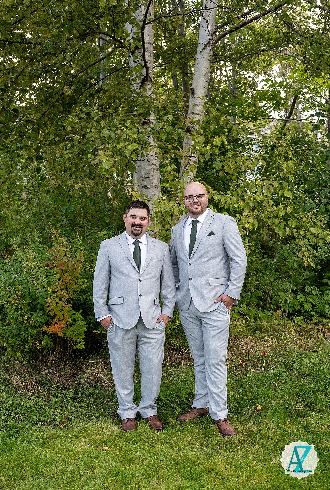 Two members of a wedding party in gray suits pose together on grass with trees in background.