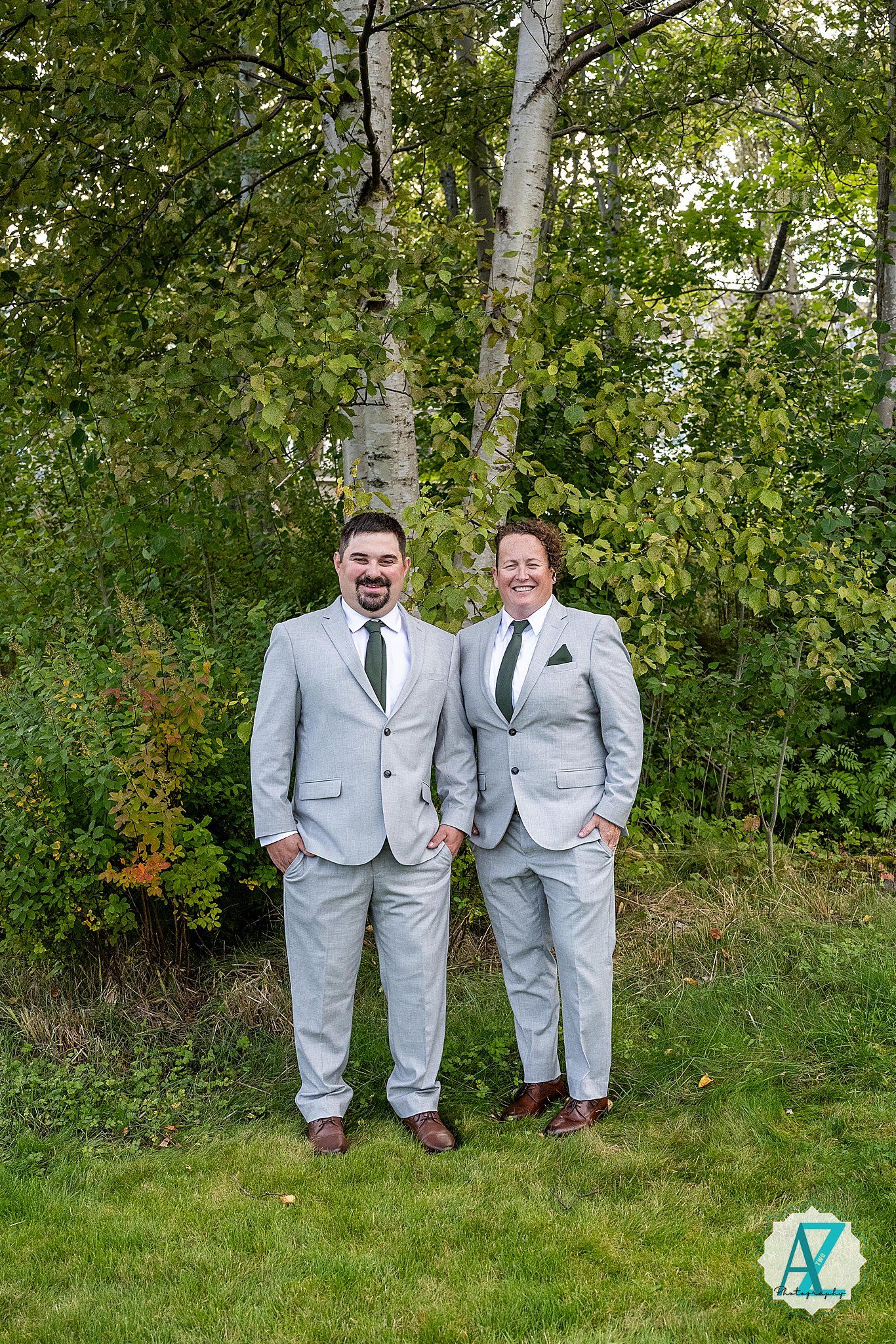 Three wedding party members in matching light gray suits standing together on grass with trees in the background.