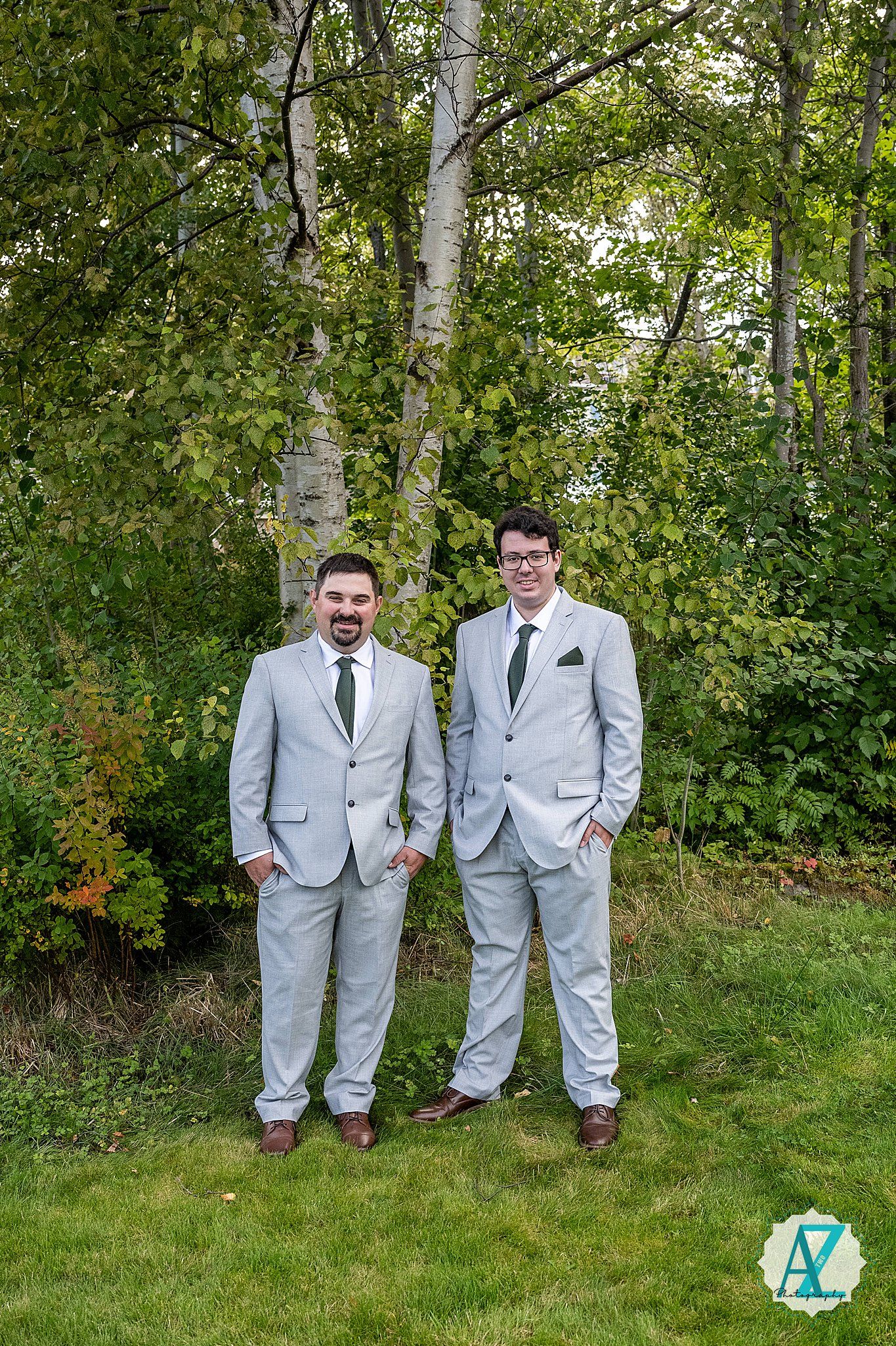 Two people in matching light gray suits stand outdoors against a backdrop of green foliage and birch trees.