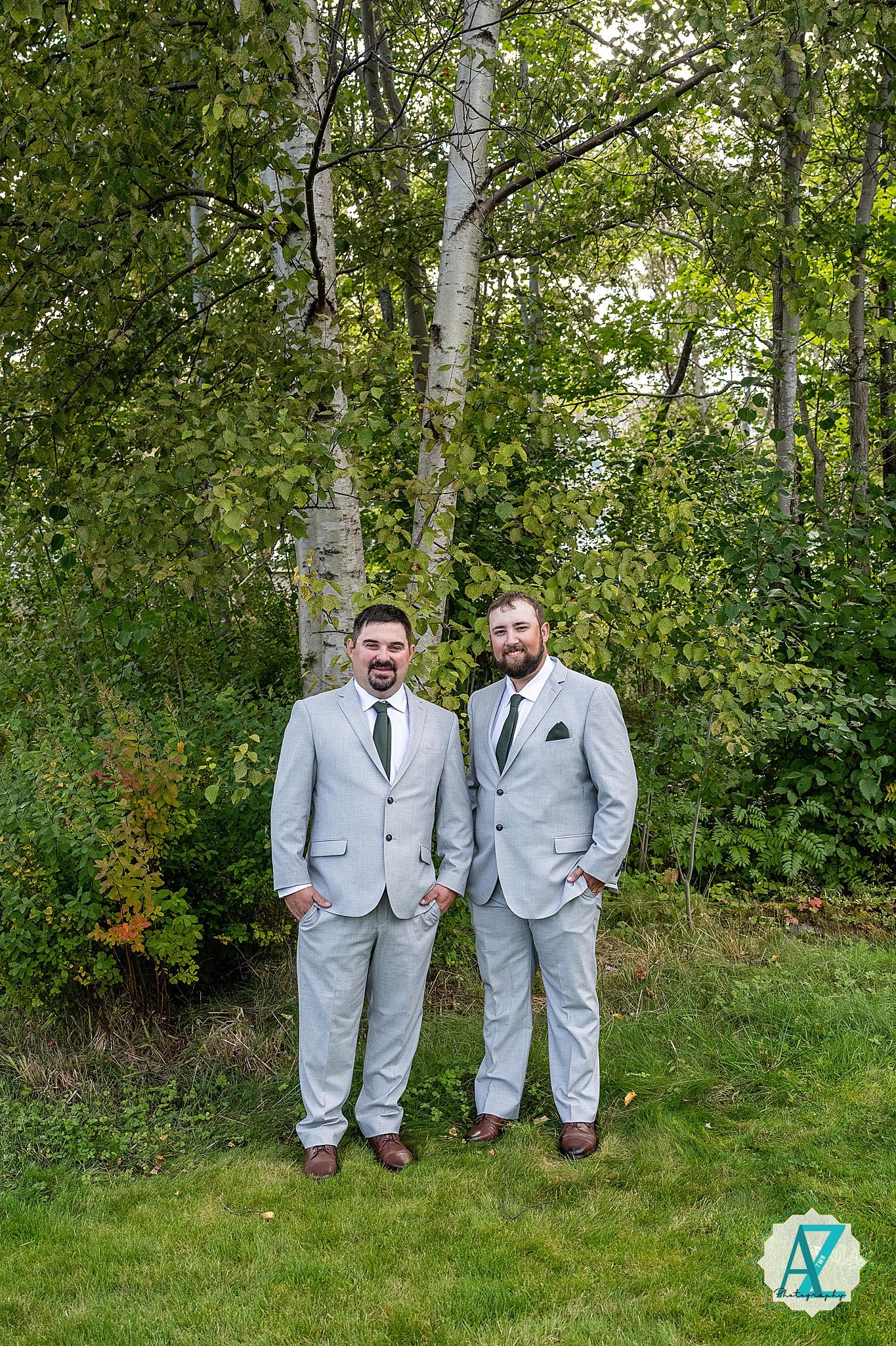 Two groomsmen in matching light gray suits stand together on grass in front of birch trees and greenery.