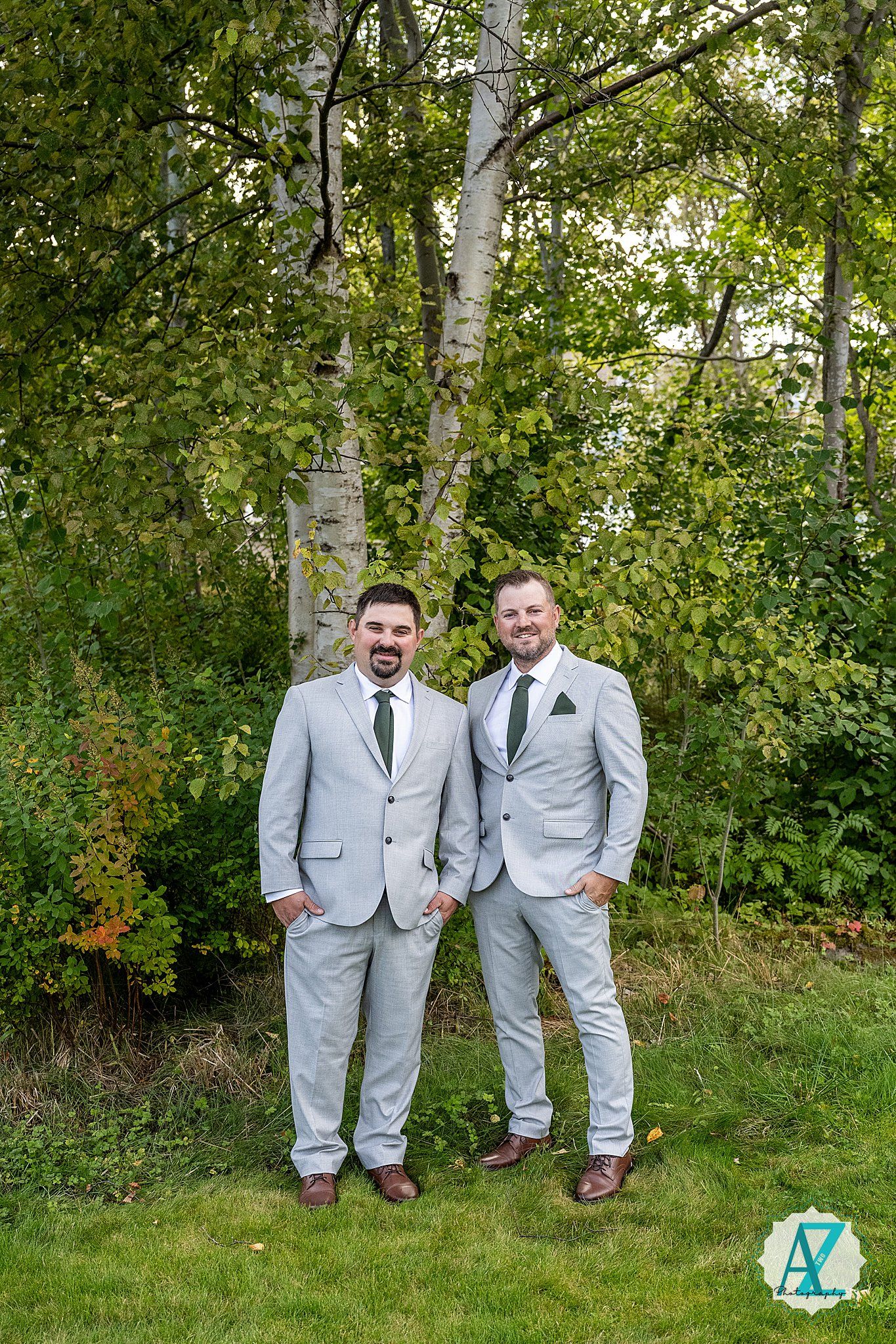 Two people in matching light gray suits pose playfully in a lush green garden setting.