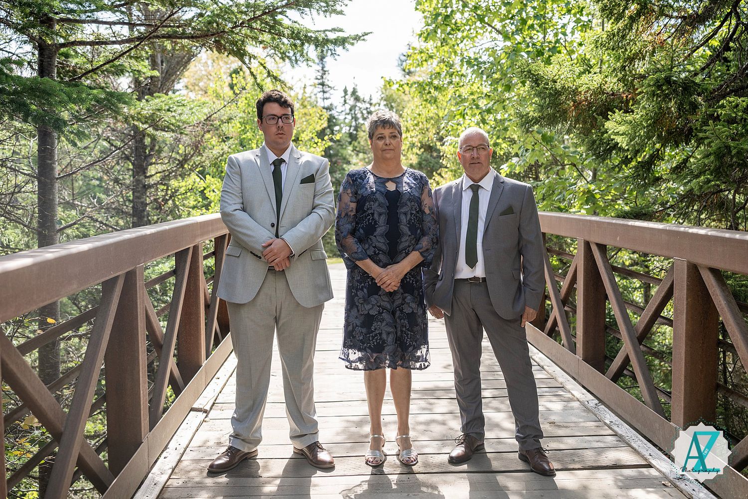 Wedding party poses on rustic wooden bridge surrounded by lush green trees during outdoor ceremony.