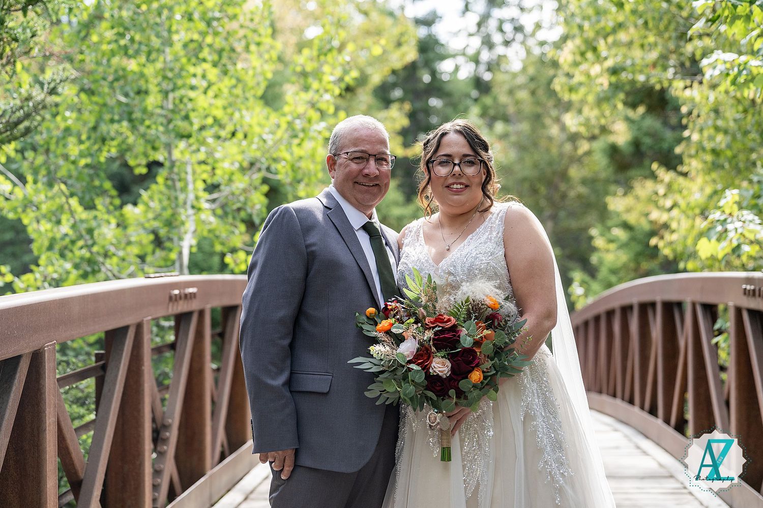 A happy couple poses together on a rustic wooden bridge surrounded by lush greenery, holding a vibrant autumn bouquet.