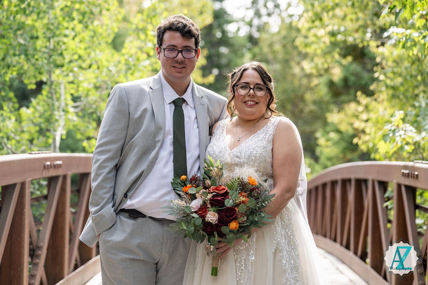 Wedding couple poses together with bouquets on wooden bridge surrounded by lush greenery in sunlight.