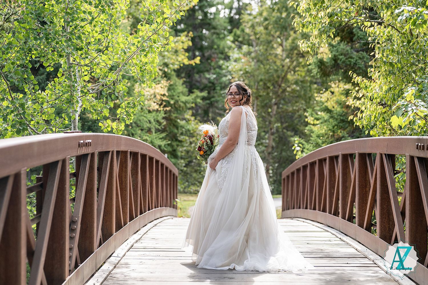 A bride in a flowing white dress poses on a rustic wooden bridge surrounded by lush green foliage on a sunny day.