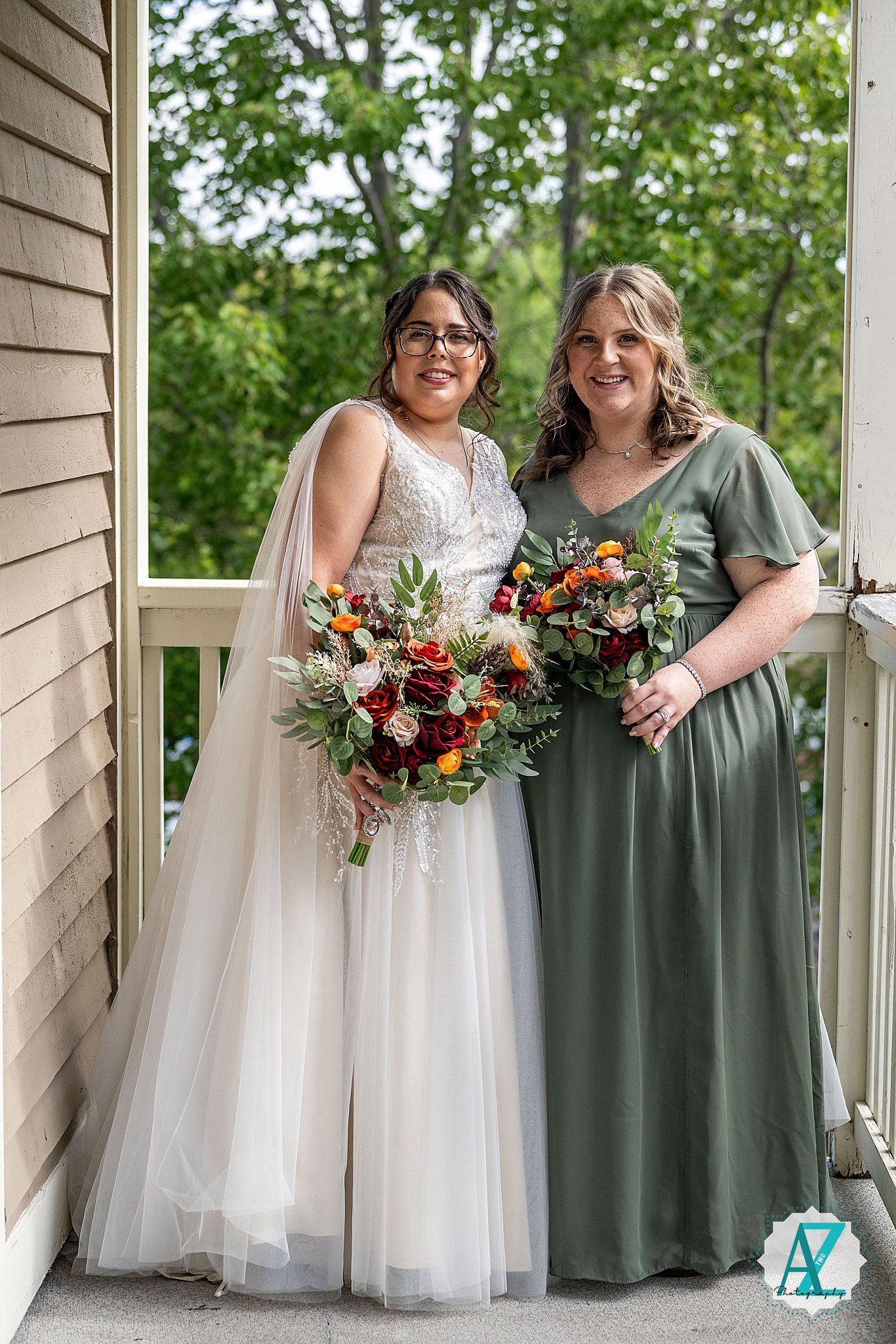 Two people stand together on a porch, one wearing a white wedding dress and one in a sage green dress, holding bouquets.