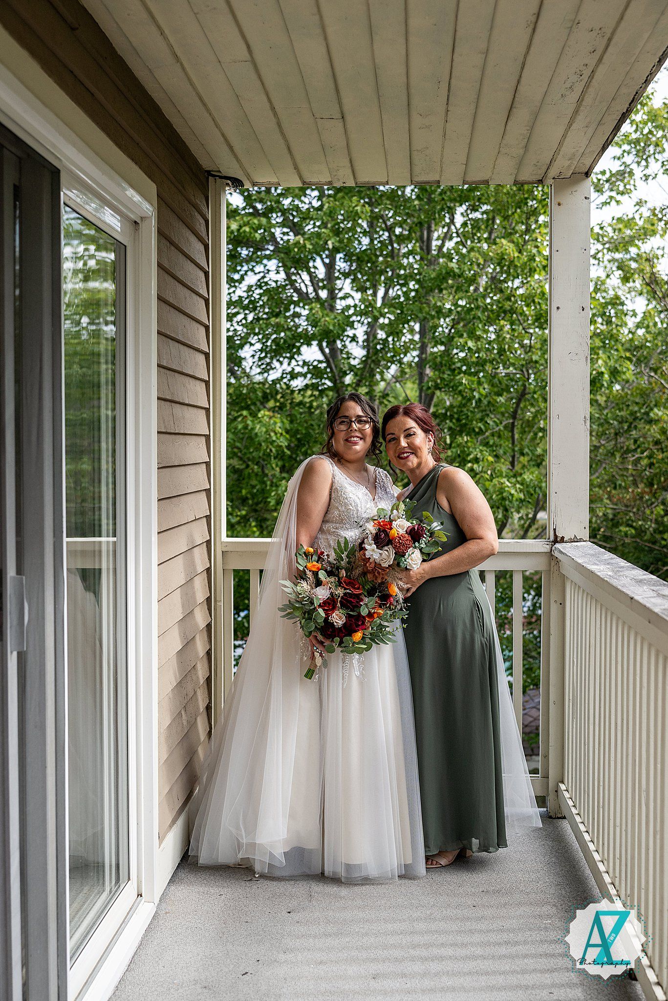 Two people in wedding attire share joyful moments on a bright wraparound porch with white railings and windows.