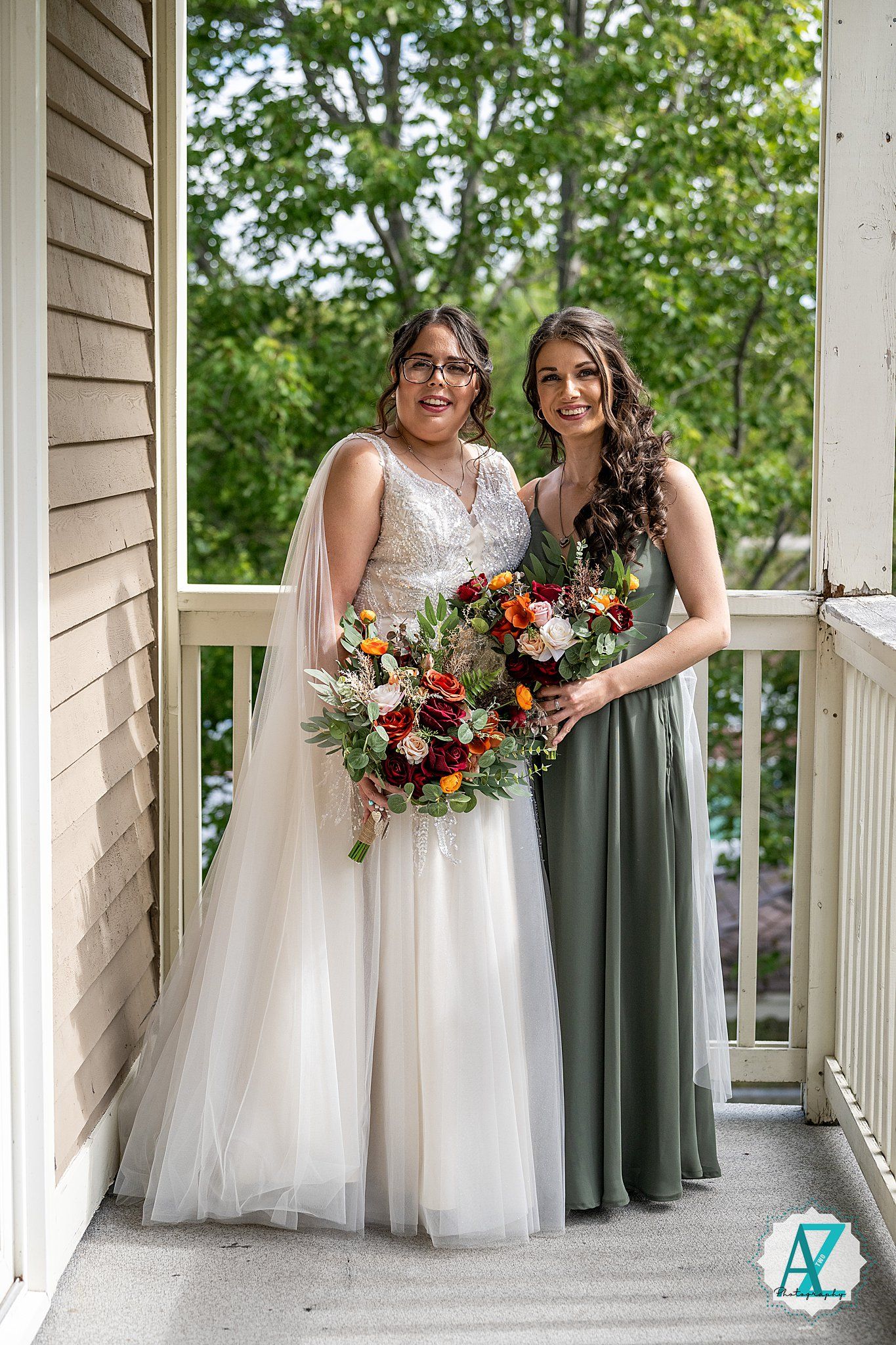 Wedding party wearing white gowns and sage green dresses pose together on a wraparound porch with vibrant flower bouquets.