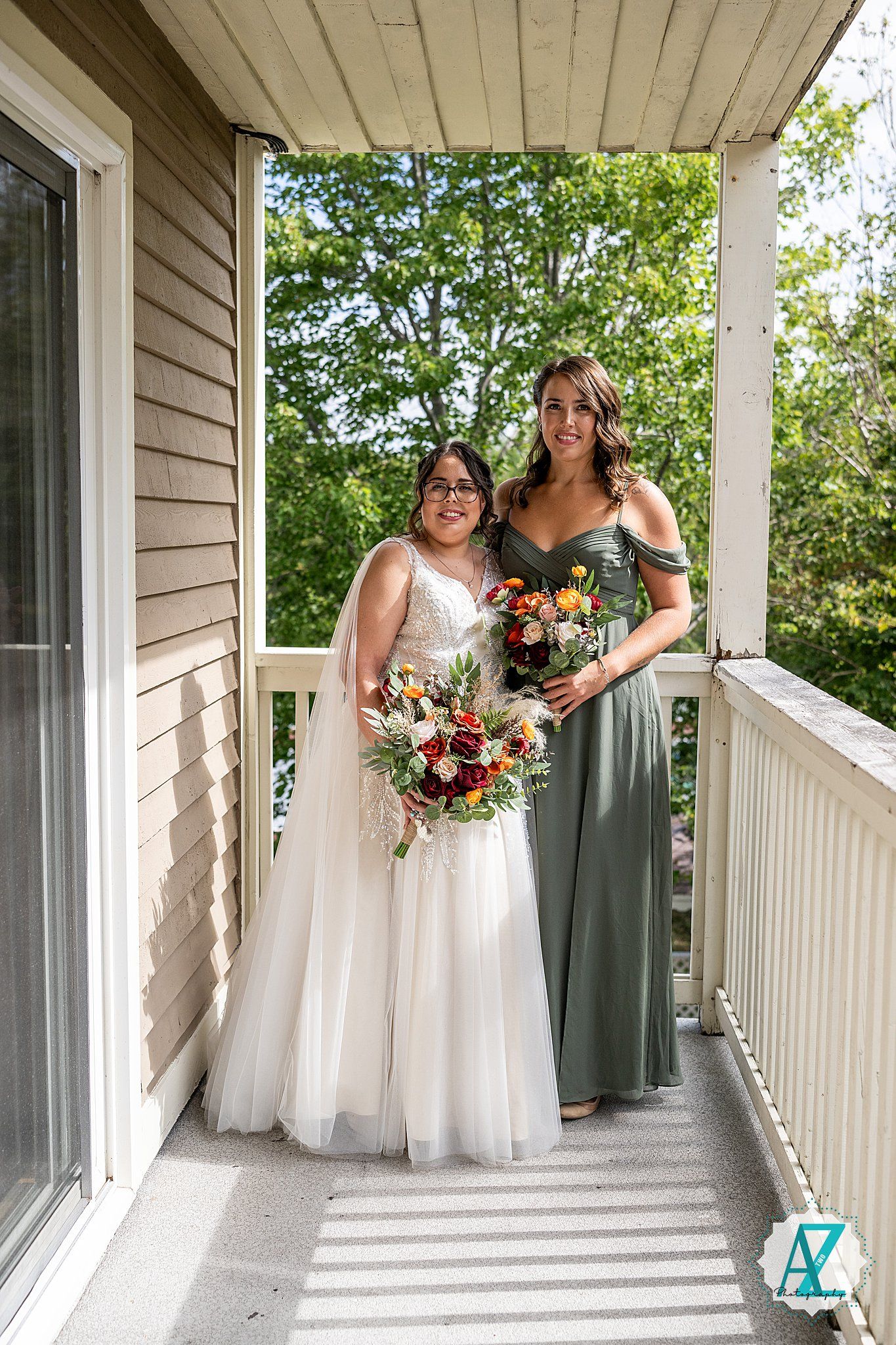 A bride in a white gown and bridesmaid in sage green dress hold colorful bouquets while posing on a covered porch.