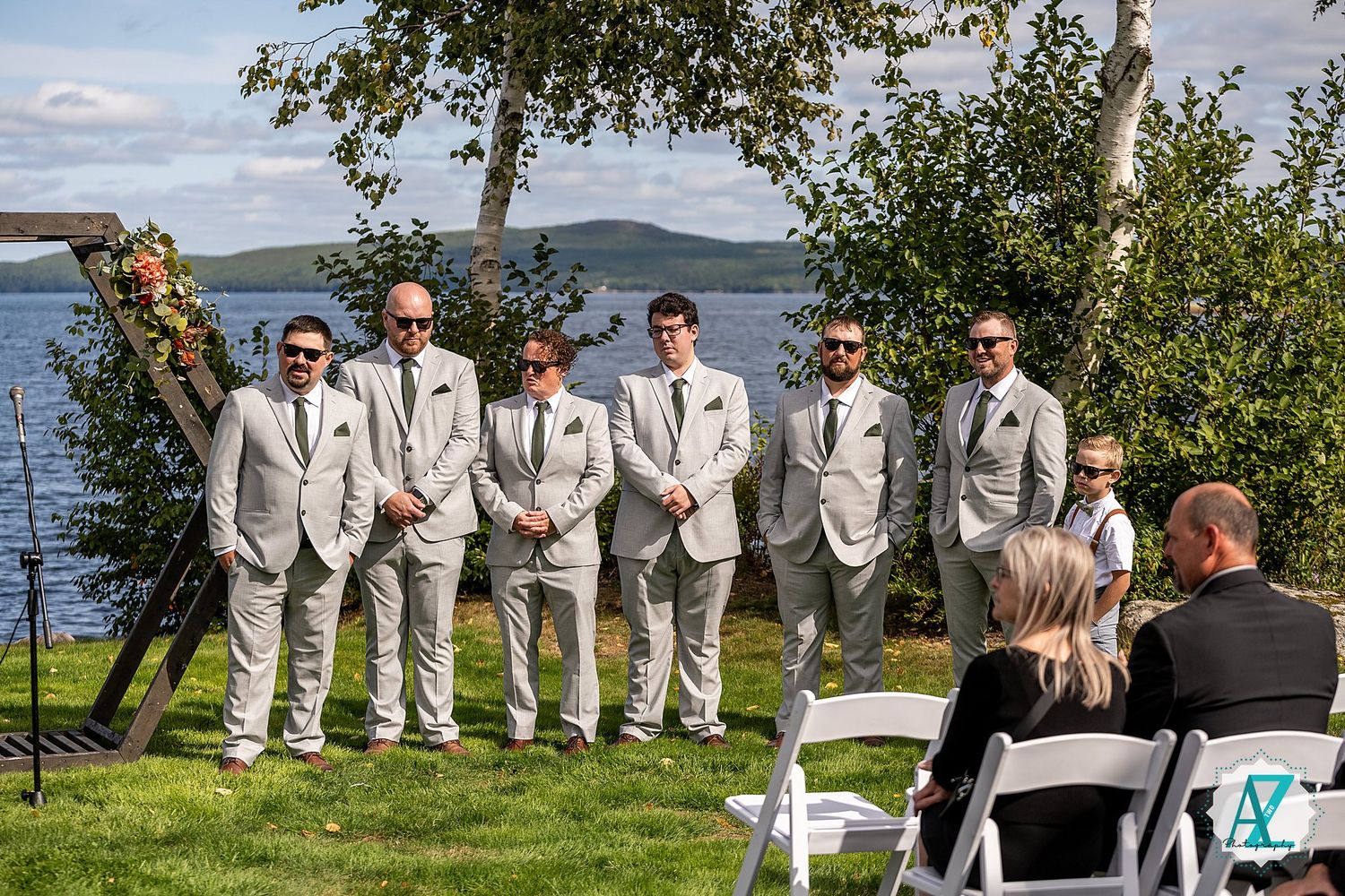 A wedding party poses outdoors in cream-colored suits against a scenic waterfront backdrop on a sunny day.