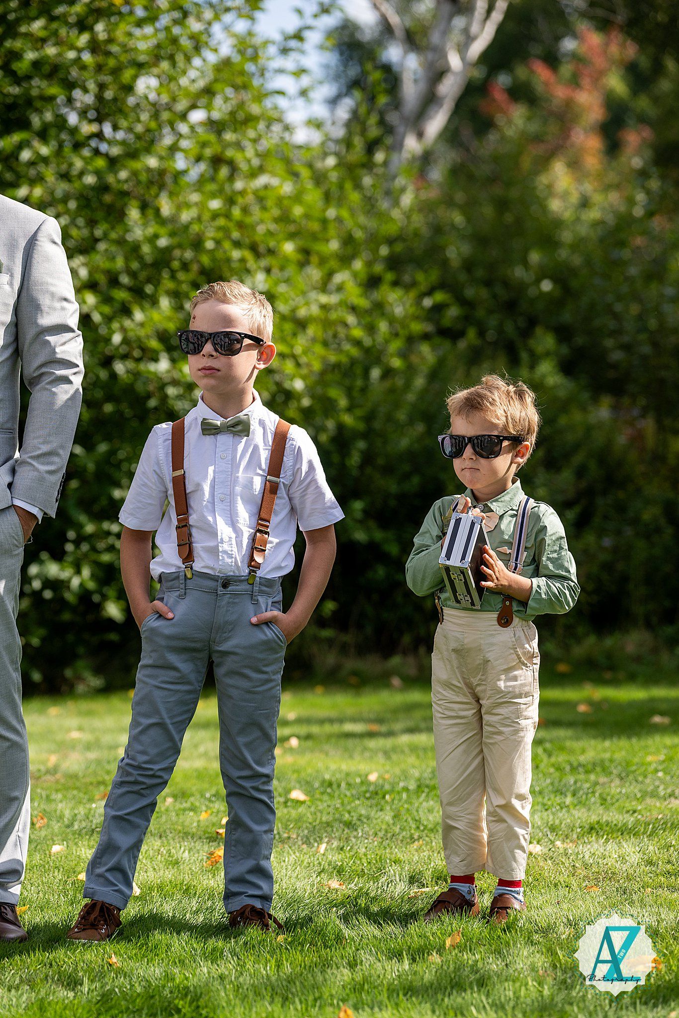 Young boys stand on grass wearing suspenders and casual attire at an outdoor gathering on a sunny day.