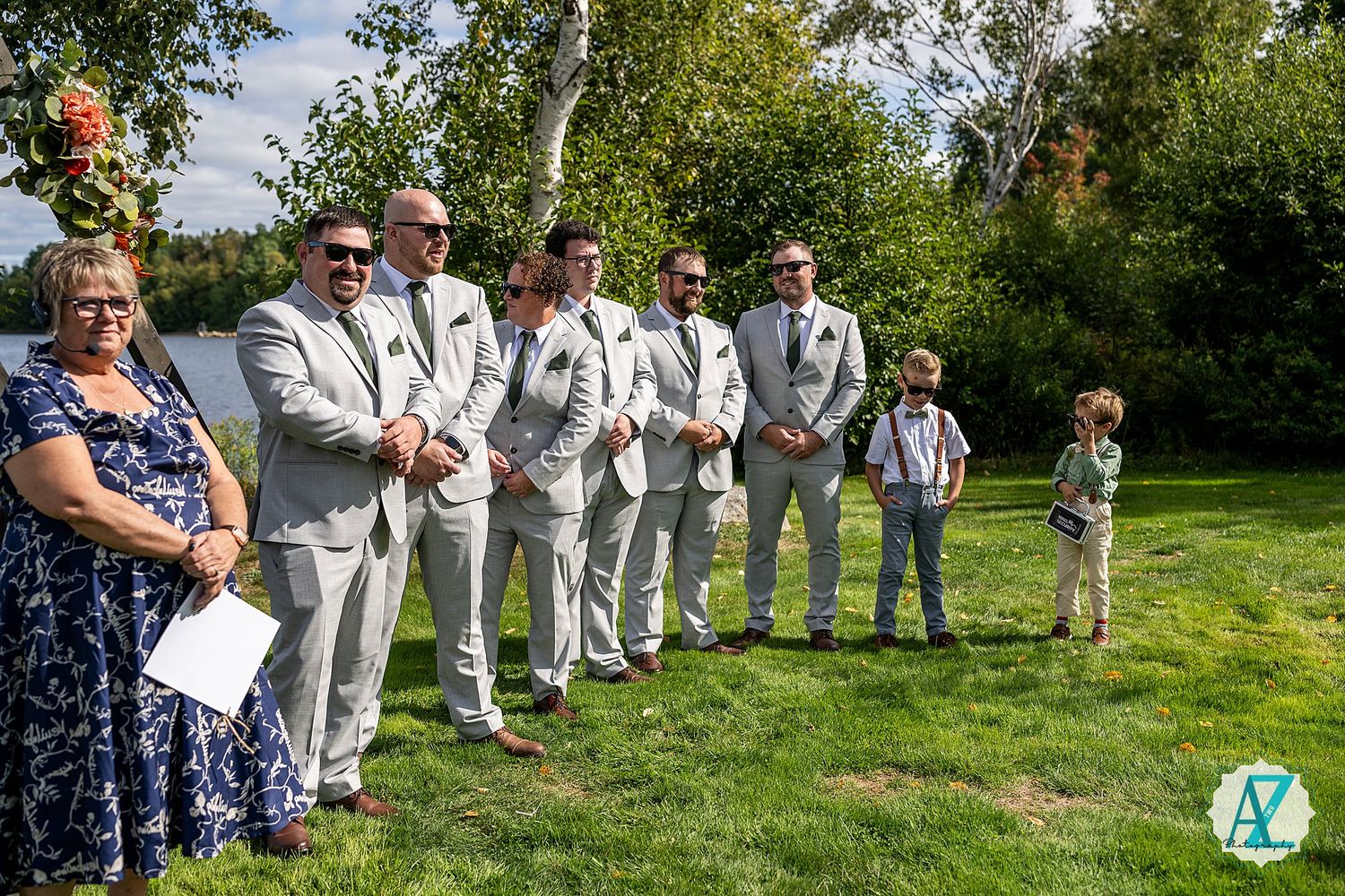 Wedding guests in light colored attire stand in line on lush green grass during an outdoor ceremony.