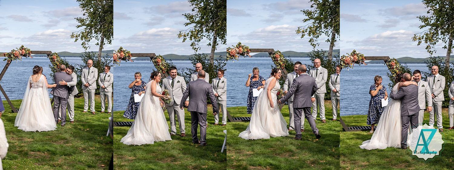 Wedding guests gather at an outdoor lakeside ceremony with trees and blue sky in the background.