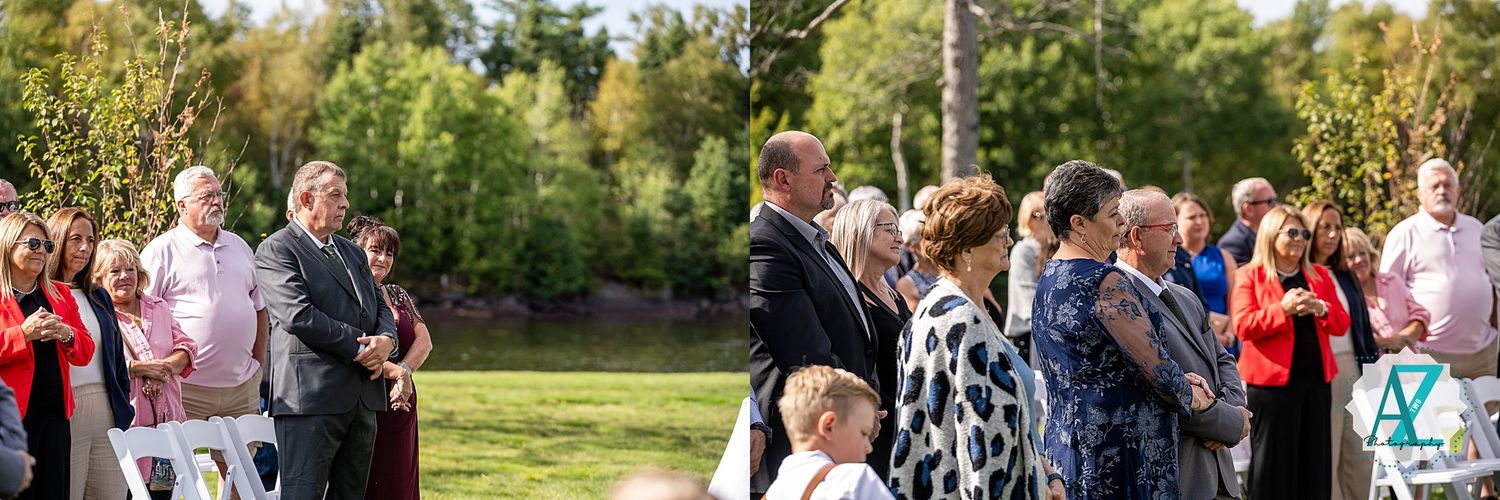 Outdoor wedding celebration with guests mingling on a grassy lawn near a lake on a sunny day.