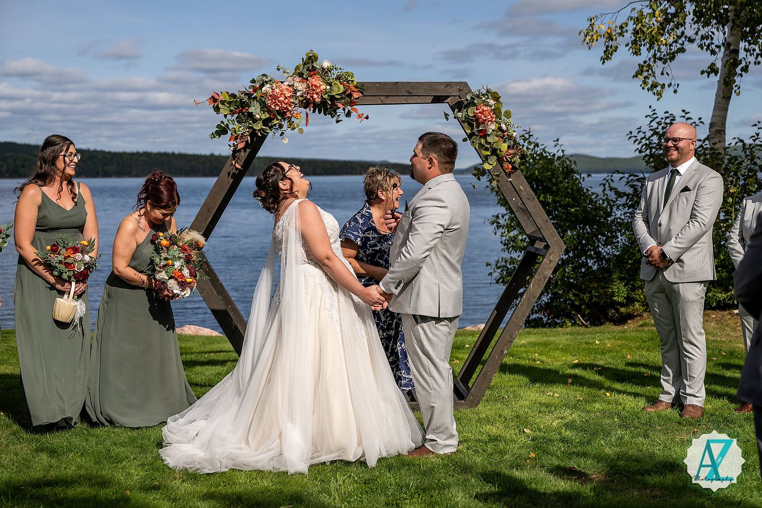 Outdoor wedding ceremony under hexagonal floral arch overlooking scenic waterfront with wedding party in sage green attire.