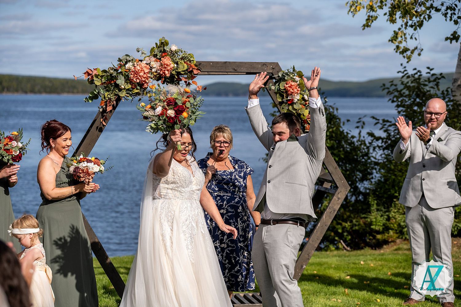 Wedding party stands together laughing during outdoor ceremony by scenic waterfront with bright summer sky.