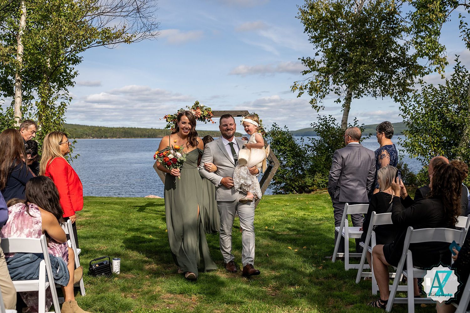 Wedding guests sitting in white chairs on a grassy lawn overlooking a serene lake at an outdoor ceremony.