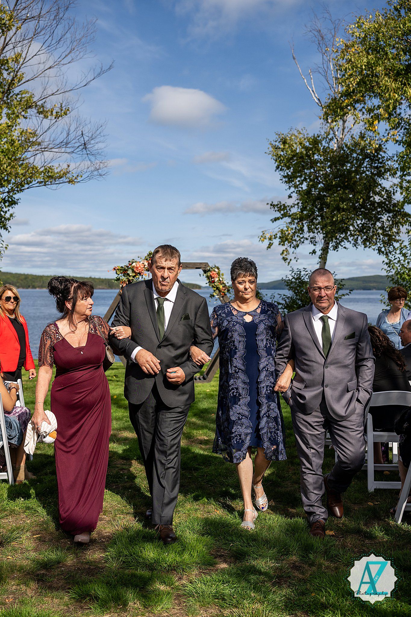Wedding party walks and laughs together on grassy hill overlooking a scenic lake on sunny afternoon.