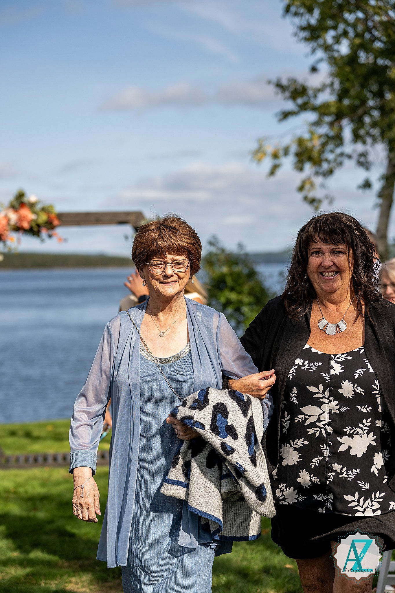 Two people stand outdoors by a waterfront on a sunny day with trees and blue sky in the background.